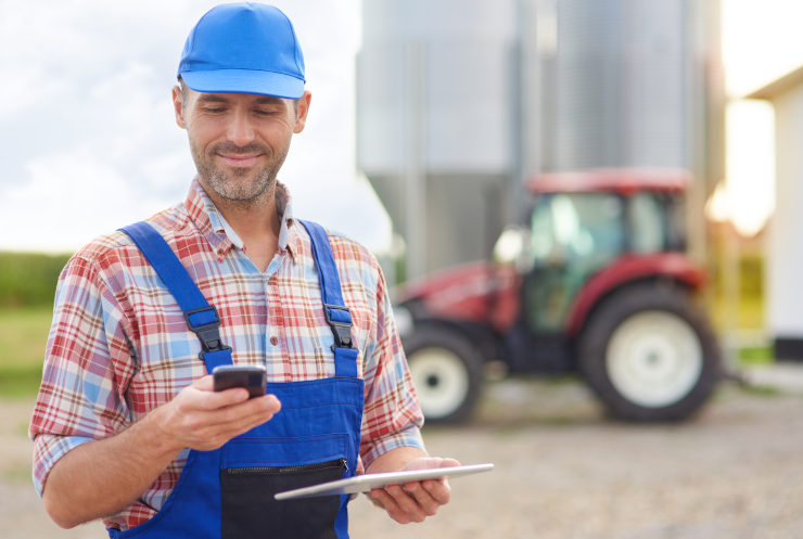 Agricultor com colete e boné azul segurando um celular e um tablet, com um trator vermelho ao fundo