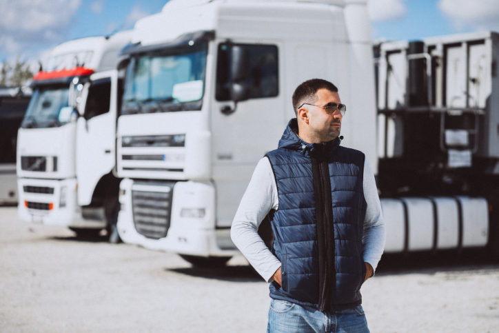 Homem com colete e óculos escuros em frente a dois caminhões